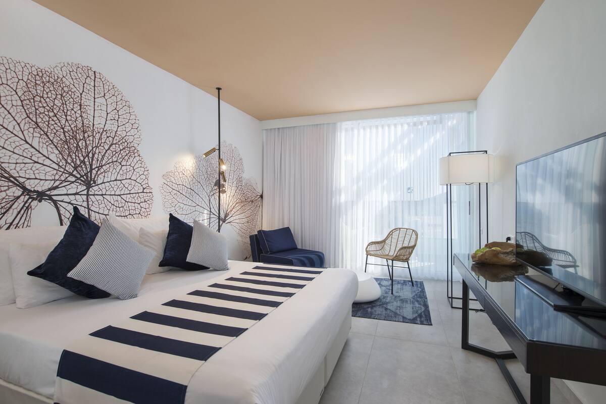 A modern hotel room with a large bed, a desk, a chair, and a large window with curtains.
