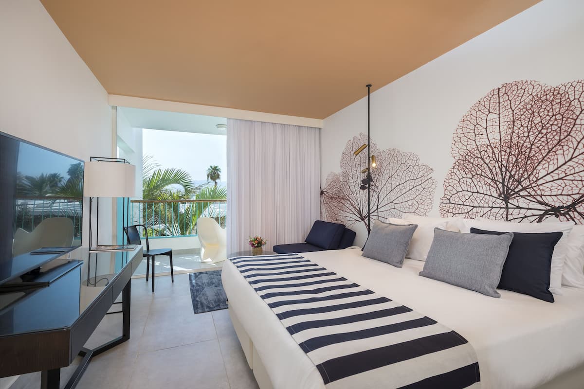 A modern hotel room with a large bed, a desk, a television, and a balcony with a view of palm trees.