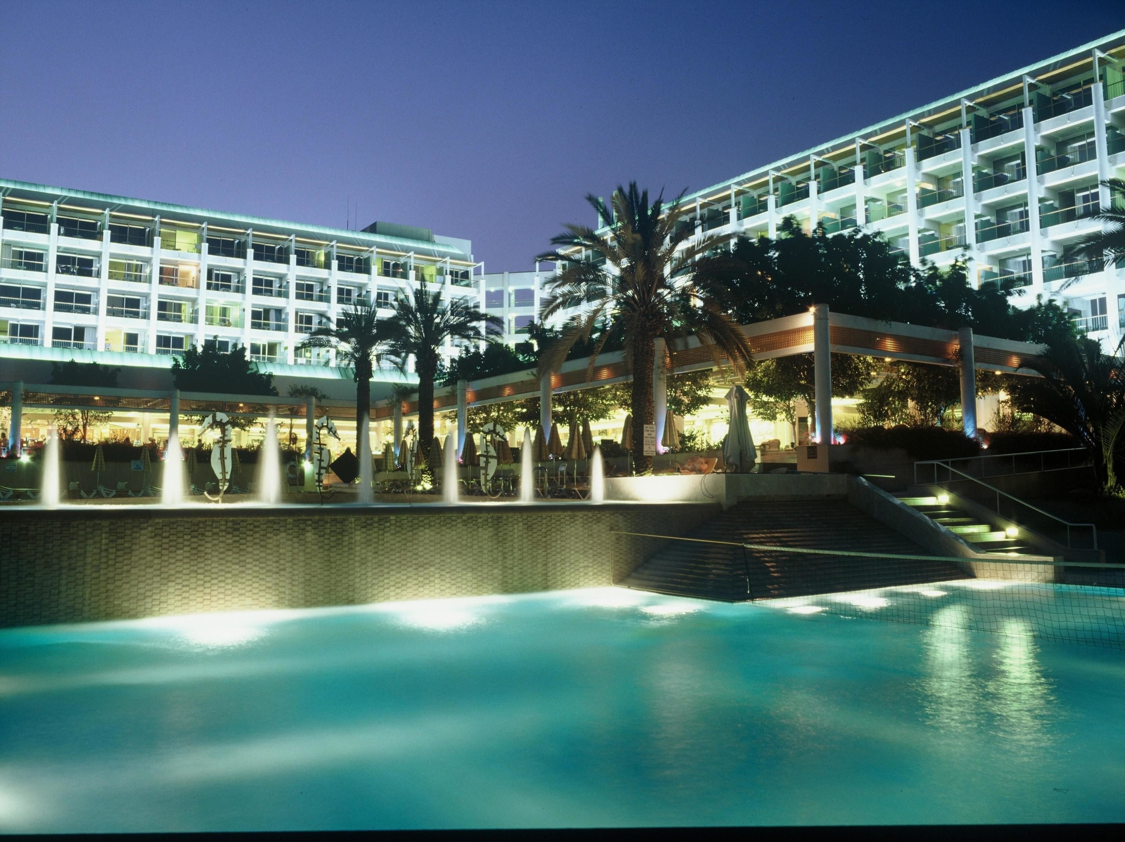 Un hotel con piscina, palmeras y una zona cubierta con sombrillas.