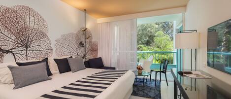 A modern hotel room with a large bed, a desk, and a balcony with a view of greenery.