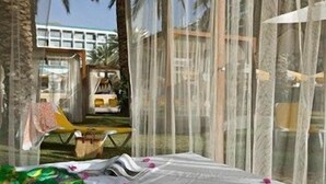 A room with a bed, a window with a view of a building and palm trees, and a chair outside. - Yam Suf by Isrotel Collection (Eilat)