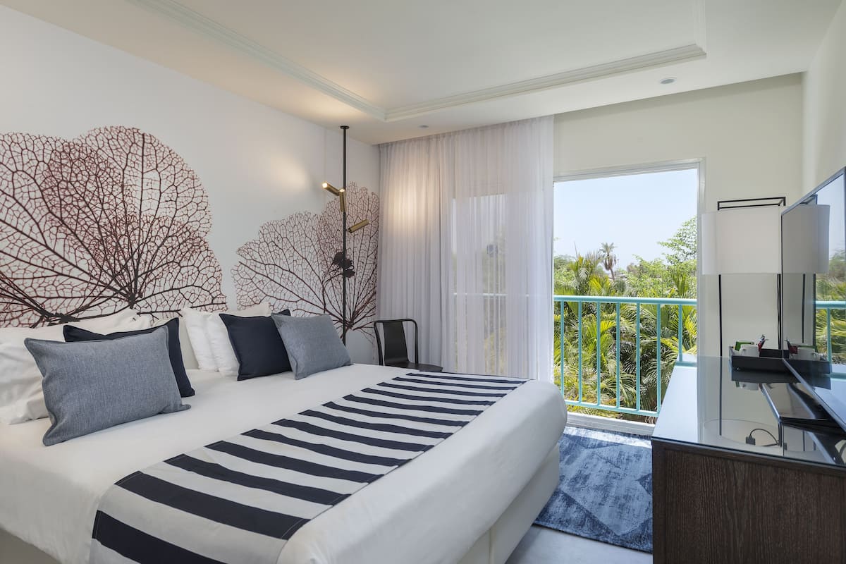 A modern hotel room with a large bed, a desk with a TV, and a balcony with a view of greenery.