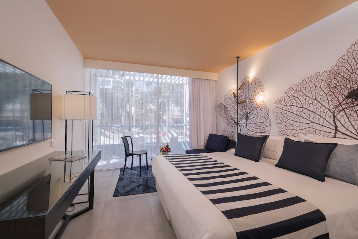 A modern hotel room with a large bed, a desk with a chair, a TV, and a large window with curtains.