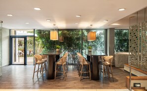 Un intérieur moderne avec une table à manger centrale entourée de chaises, de grandes fenêtres offrant une vue sur la verdure et un éclairage encastré au plafond.