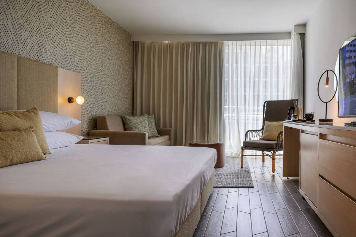 A hotel room with a large bed, a desk, a chair, and a view of the city through the window.