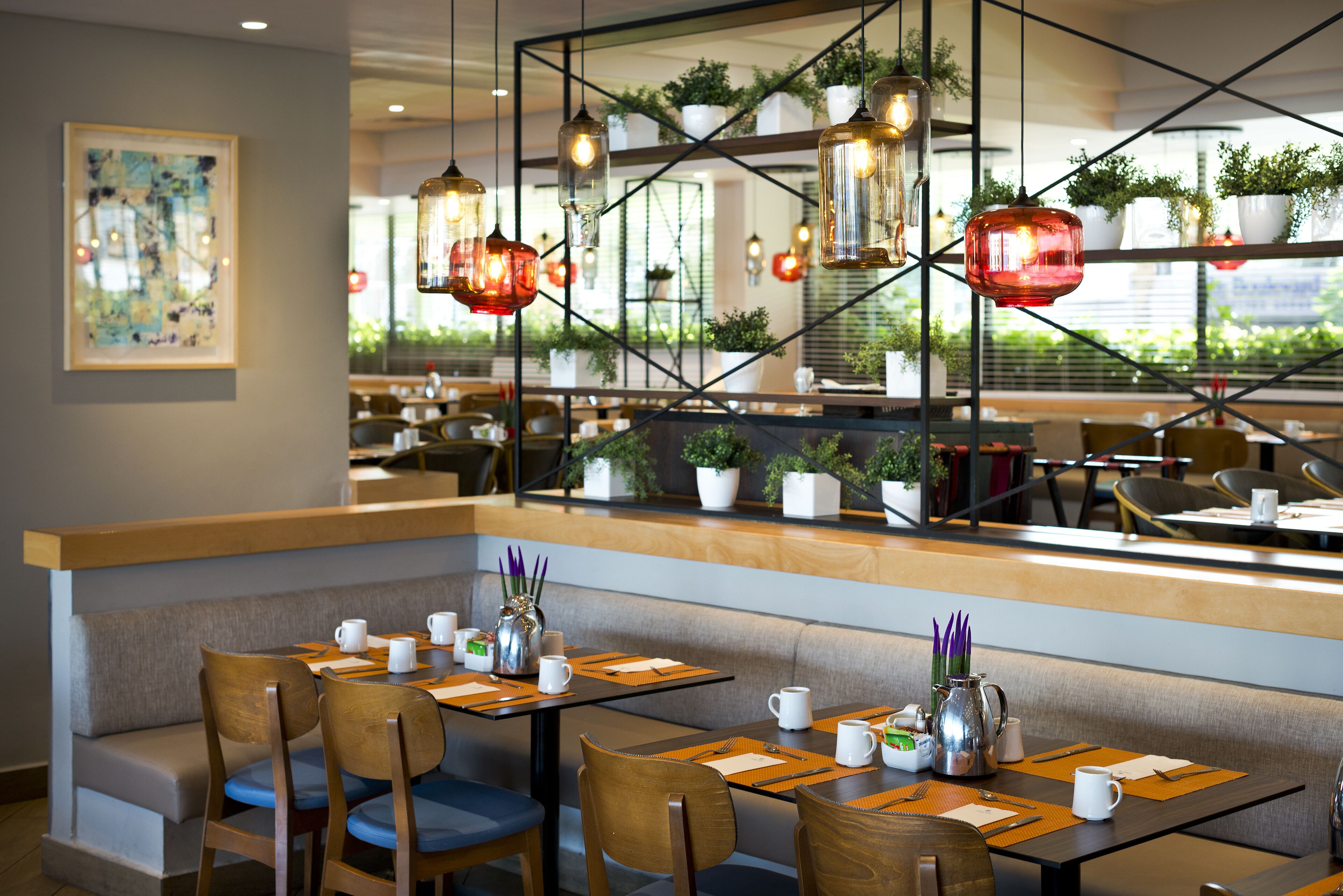 Um restaurante moderno com mesas de madeira, assentos acolchoados, luminárias pendentes e plantas em vasos.