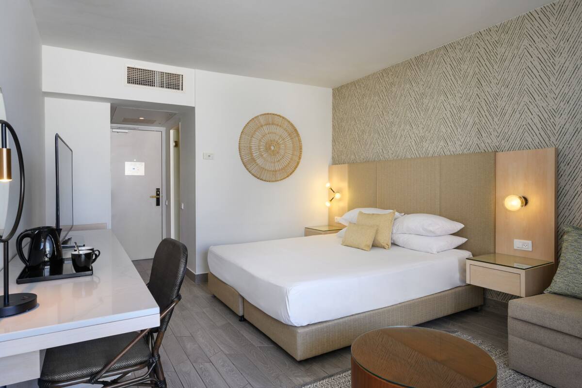 A hotel room with a bed, bedside tables, a desk with a chair, a coffee maker, and a wall-mounted decorative item.