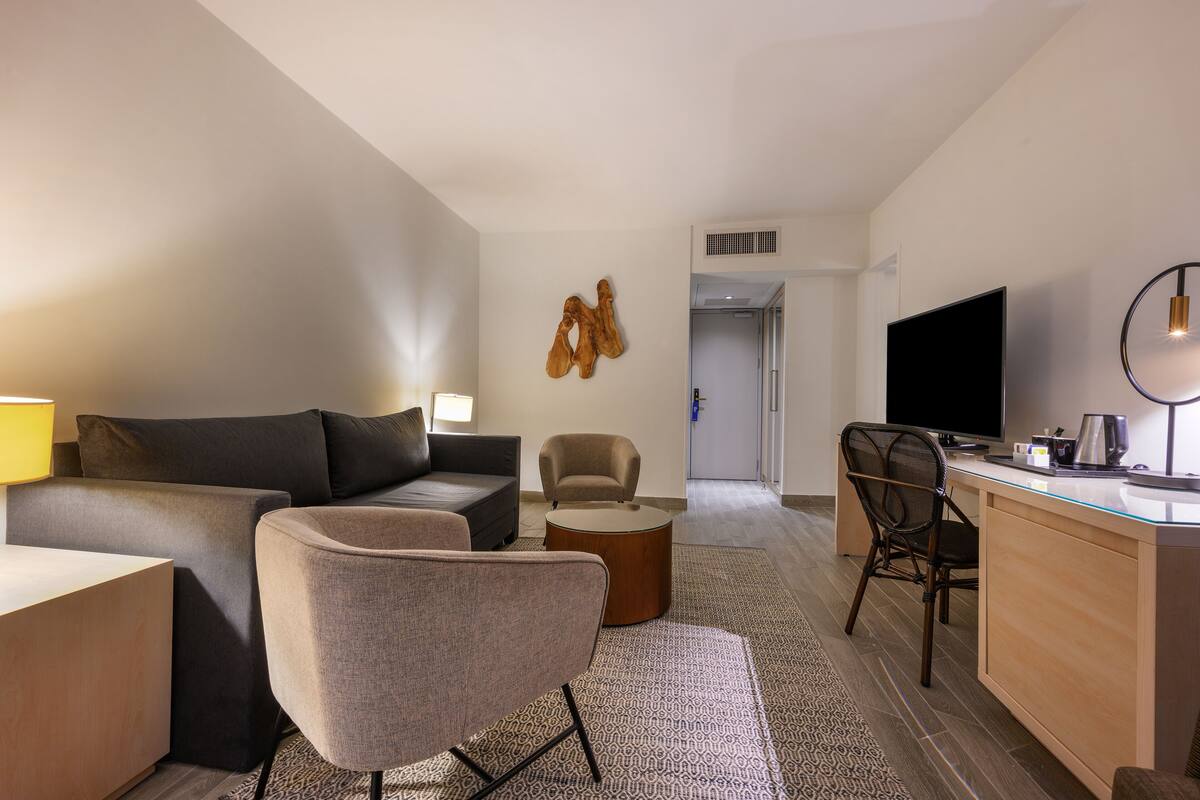 A modern hotel room with a sofa, armchair, desk, and TV.