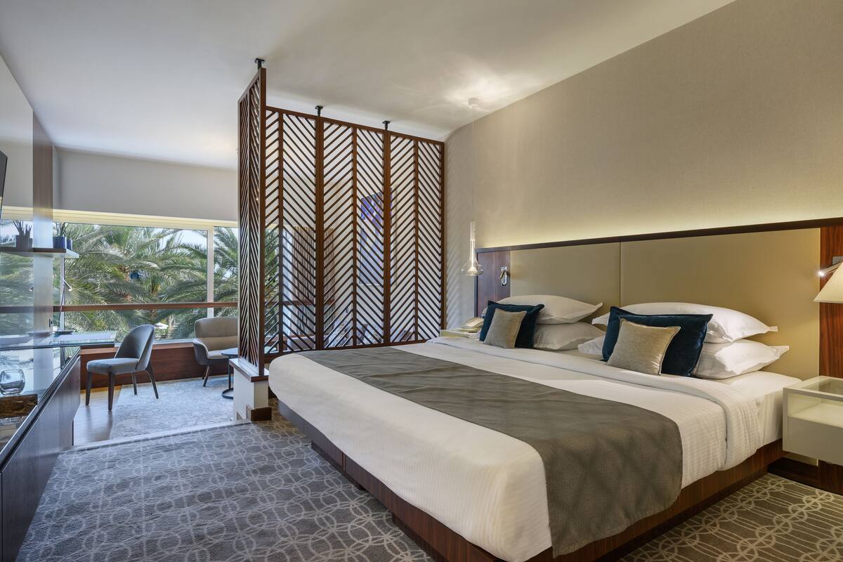 Une chambre d’hôtel avec un grand lit, un bureau avec une chaise, une vue sur des palmiers et un paravent décoratif en bois.