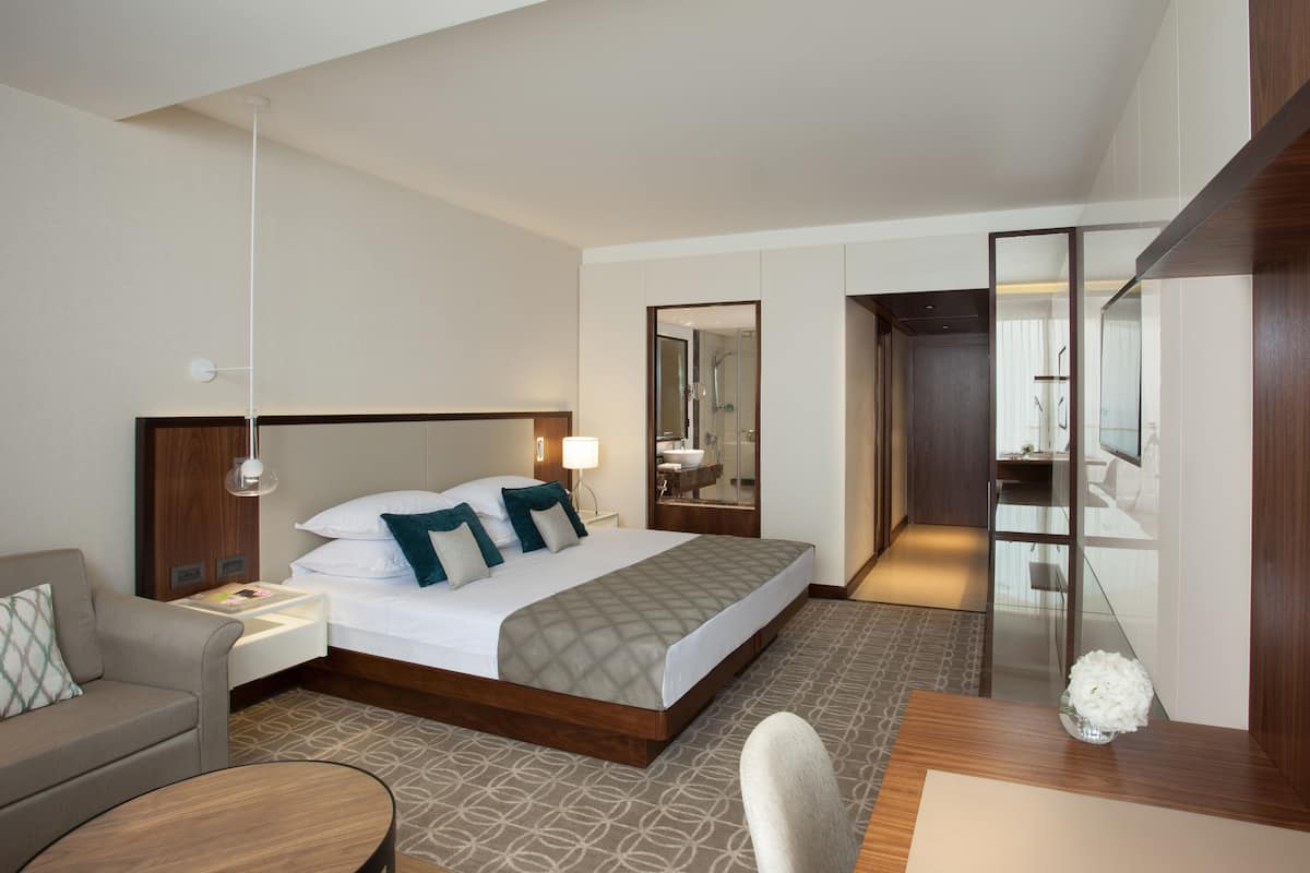 Une chambre d’hôtel moderne dotée d’un grand lit, d’un bureau, d’une chaise, d’un canapé et d’une salle de bain visible à travers une porte en verre.