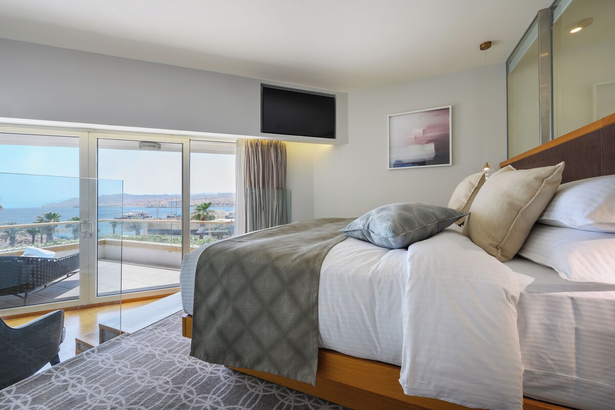 A modern bedroom with a large bed, a TV mounted on the wall, and a balcony with a view of the sea.
