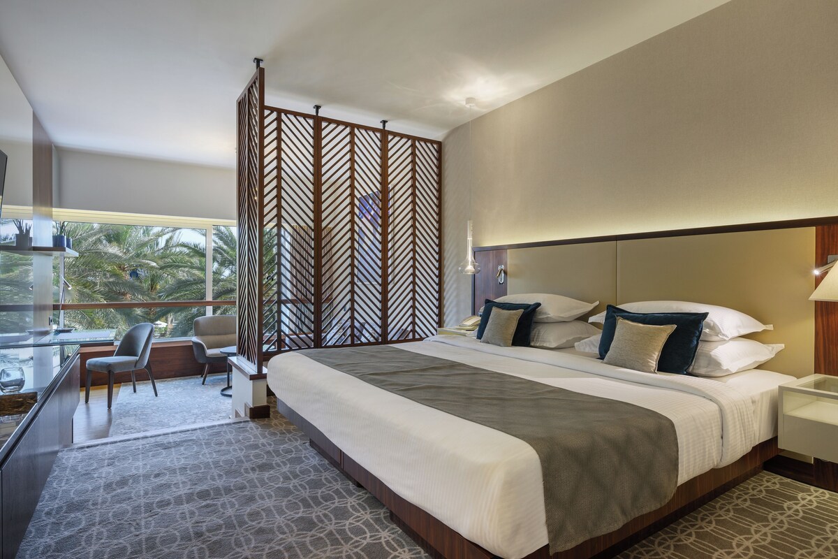 A hotel room with a large bed, a desk with a chair, a view of palm trees, and a decorative wooden partition.