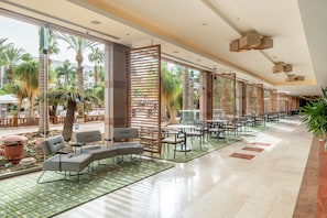 A spacious indoor area with a long row of tables and chairs, large windows with a view of palm trees, and a tiled floor. - Royal Garden by Isrotel Collection (Eilat)