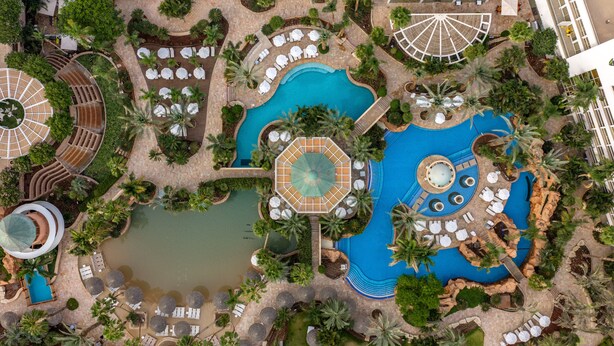 Vista aérea de un complejo turístico con varias piscinas, tumbonas y palmeras.