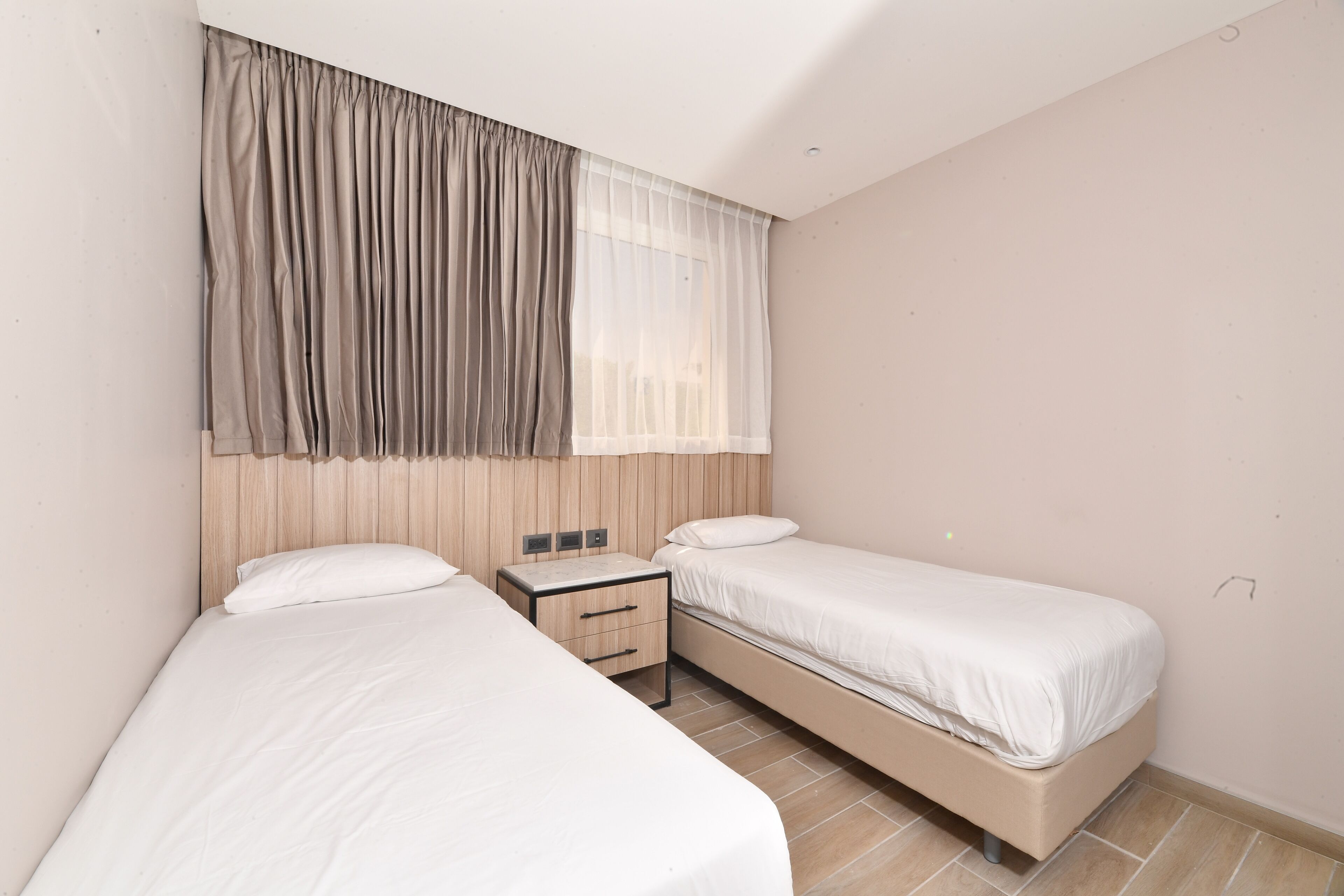 Une chambre avec deux lits simples, une table de chevet, une fenêtre avec des rideaux légers et une tête de lit en bois.