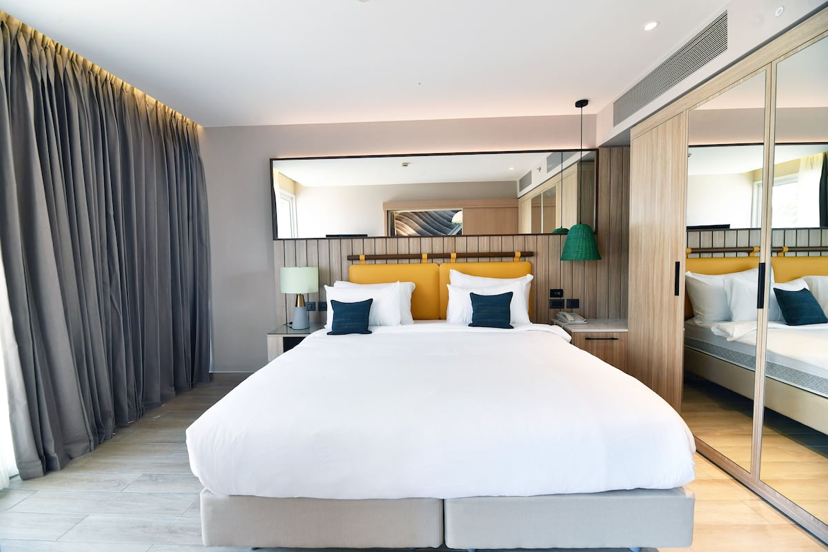 A modern hotel room with a large bed, a bedside table with a lamp, a mirror, and a view of the city through the window.