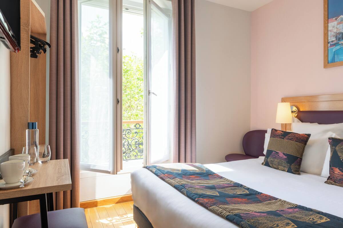 Habitación de hotel con cama, escritorio, silla, lámpara y vistas al exterior.
