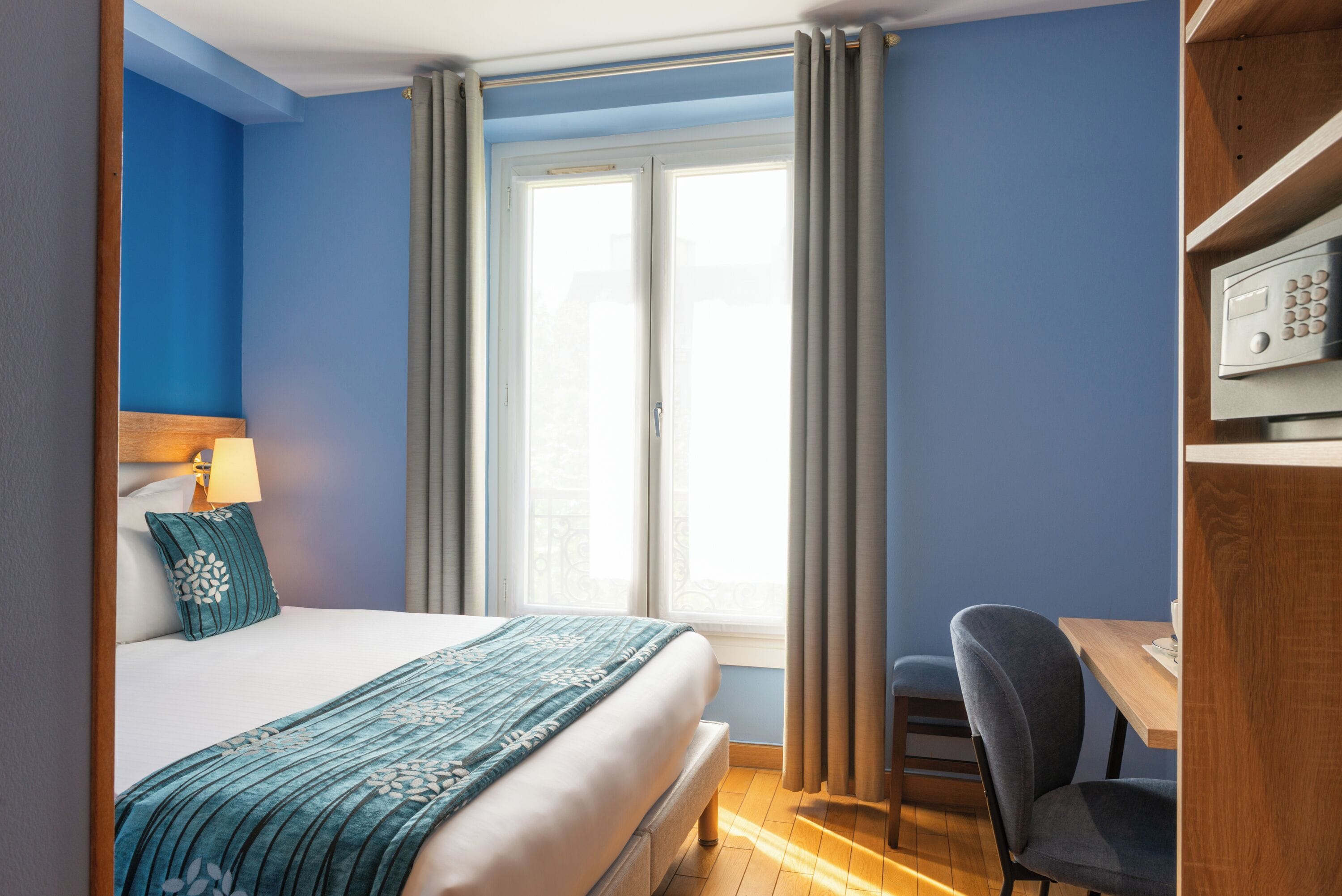 A hotel room with a bed, a desk, a chair, a lamp, and a window with curtains.
