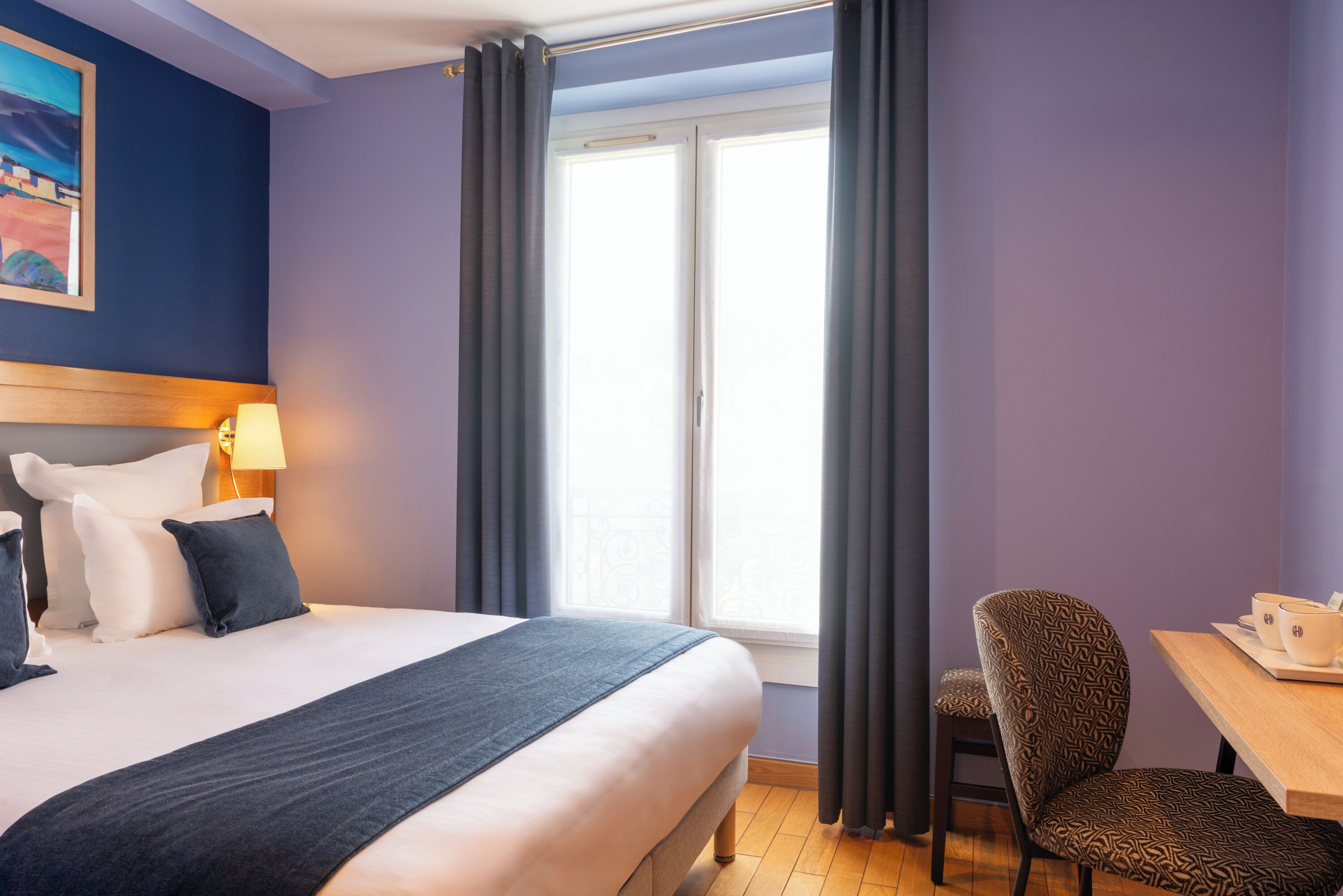A hotel room with a bed, a chair, a table, a lamp, and a window with curtains.