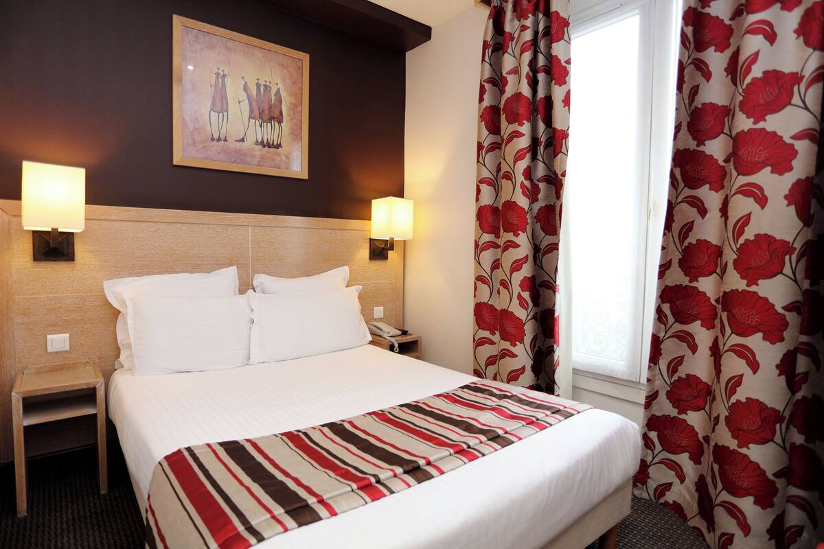 Hotelkamer met een bed, twee bedlampen, een nachtkastje, een schilderij aan de muur en een raam met bloemen gordijnen.