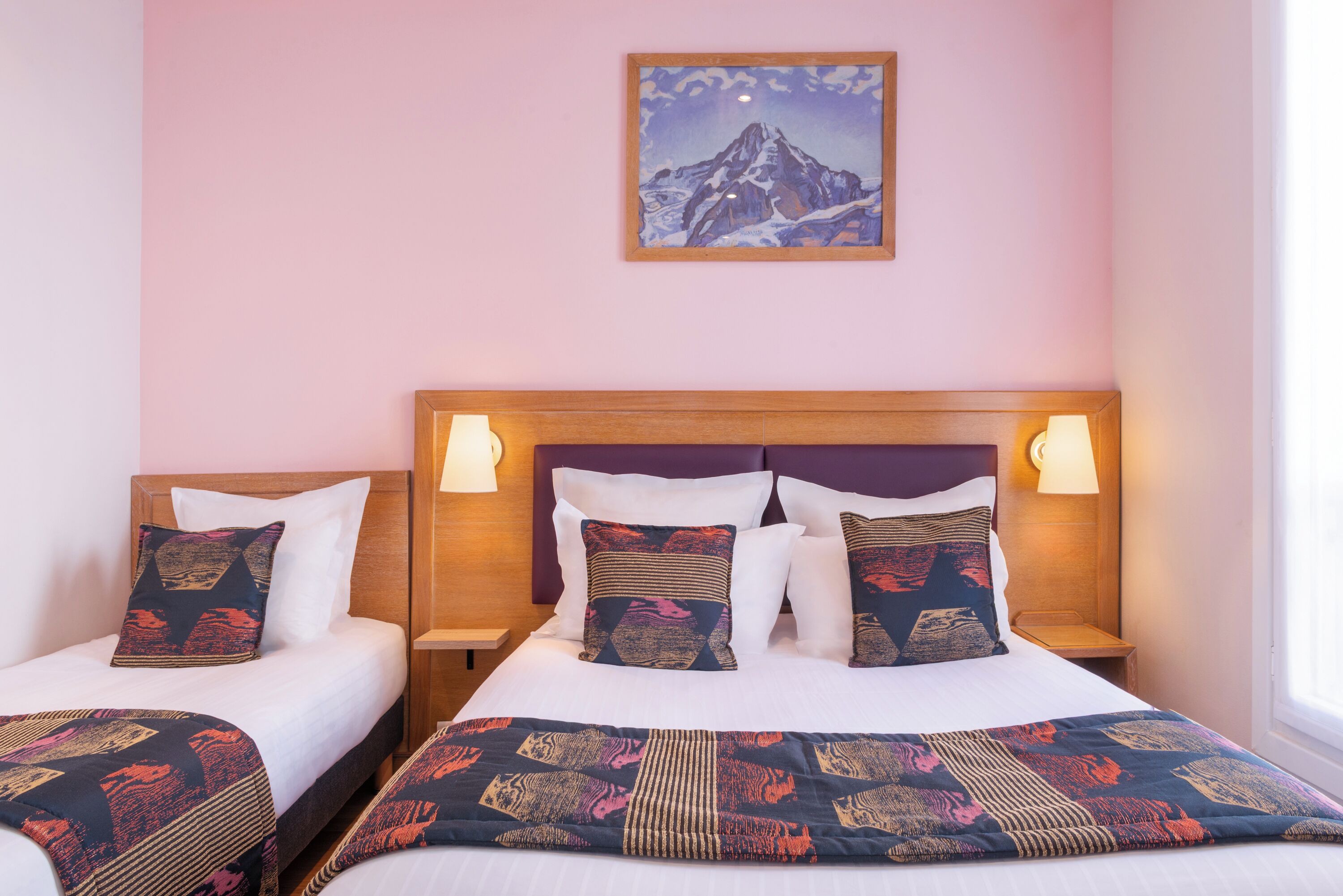 A hotel room with two beds, each with patterned bedspreads and pillows, a wooden headboard, bedside lamps, and a framed picture on the wall.