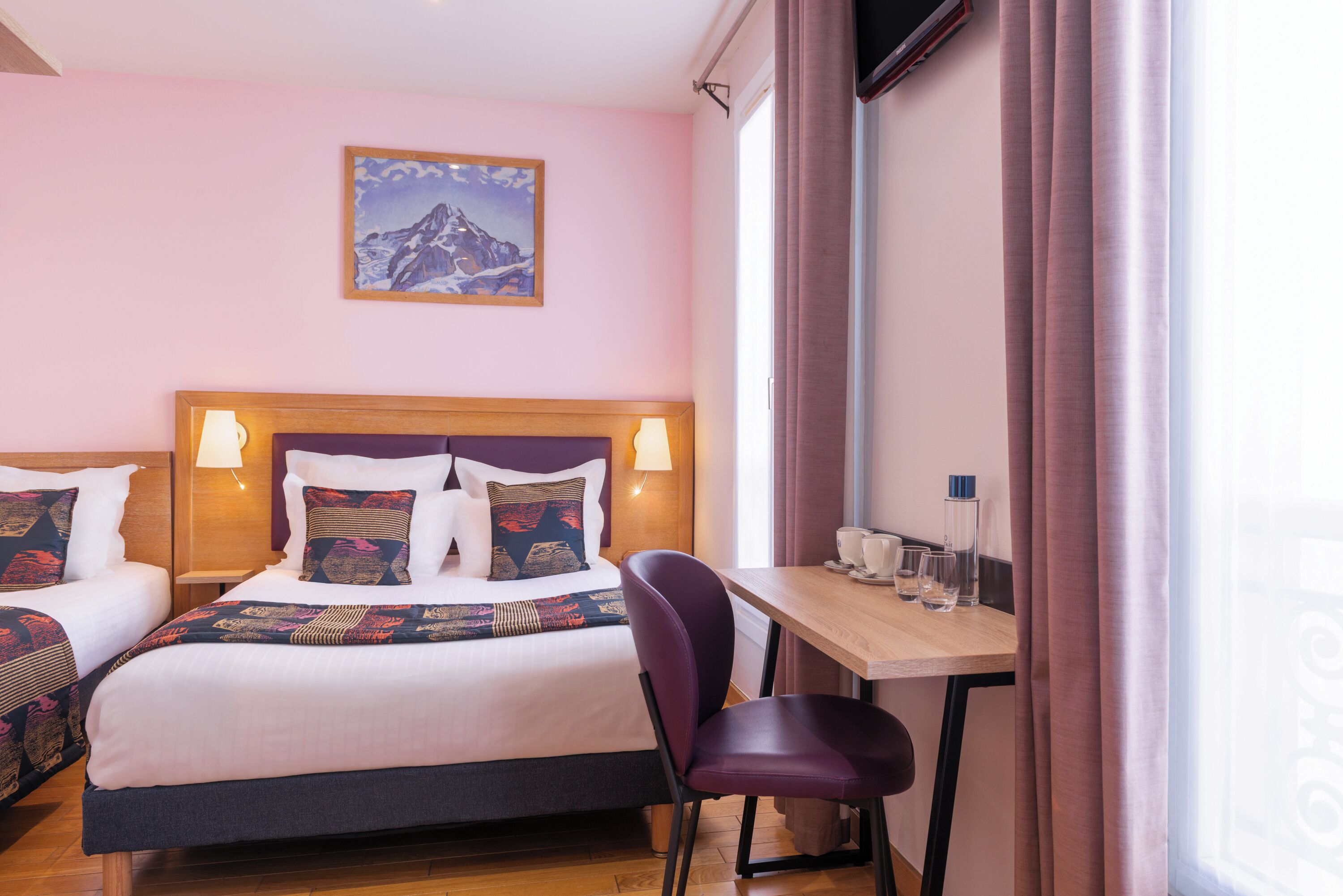 A hotel room with two beds, a wooden headboard, a desk with a chair, and a mountain painting on the wall.