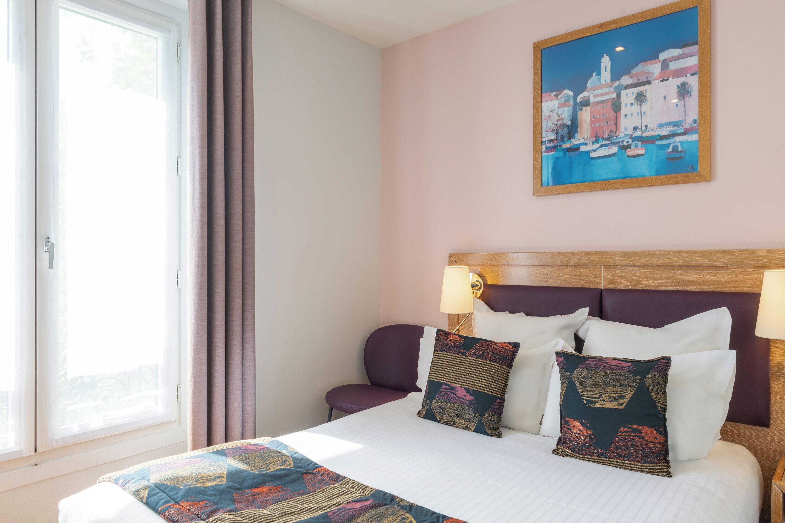 A hotel room with a bed, a nightstand, a lamp, a window with curtains, and a painting on the wall.