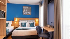 A hotel room with two beds, a desk, a chair, a framed picture on the wall, and a window with curtains. - Grand Hotel Dore (Paris)