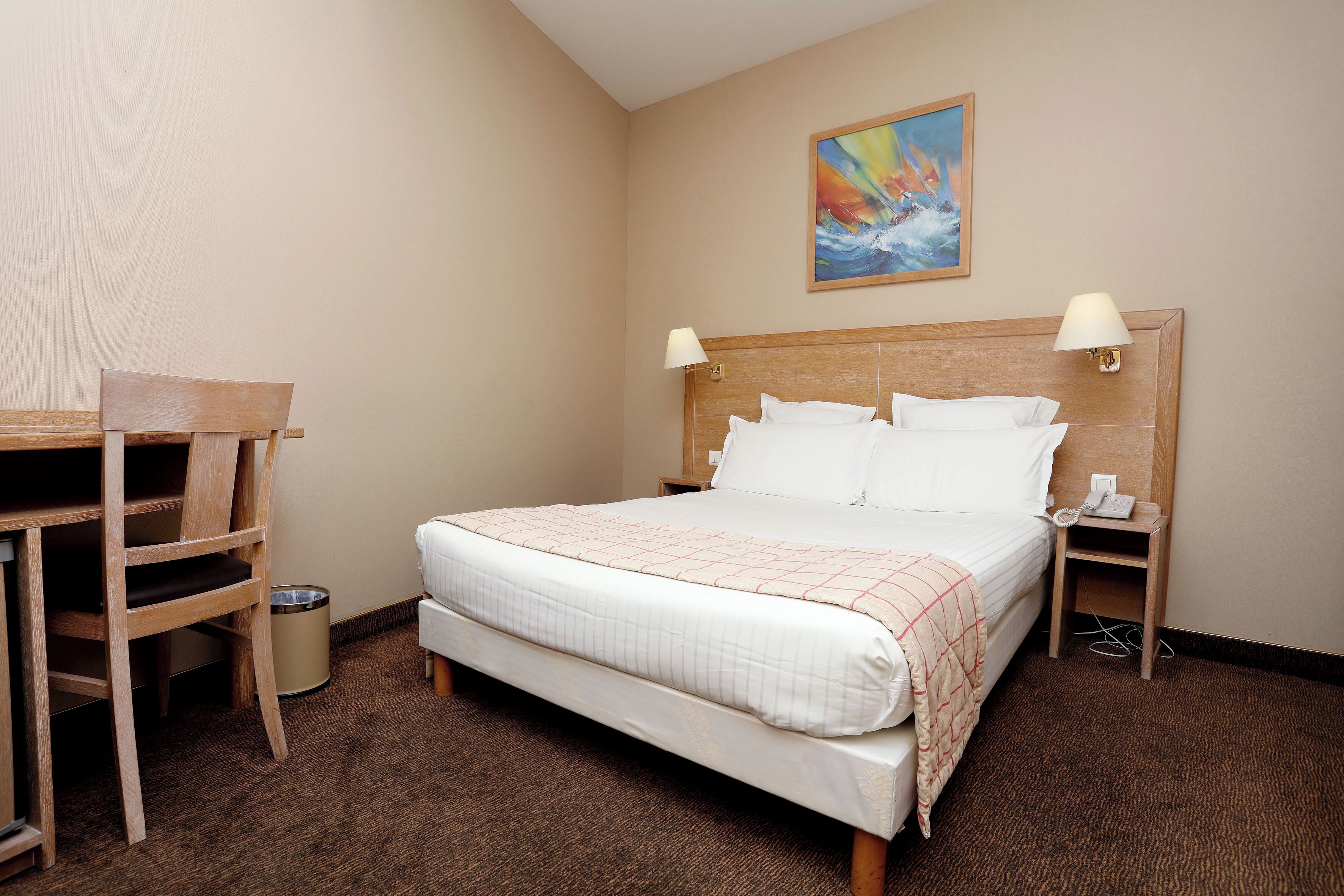 A hotel room with a bed, a desk, a chair, a trash bin, and a painting on the wall.