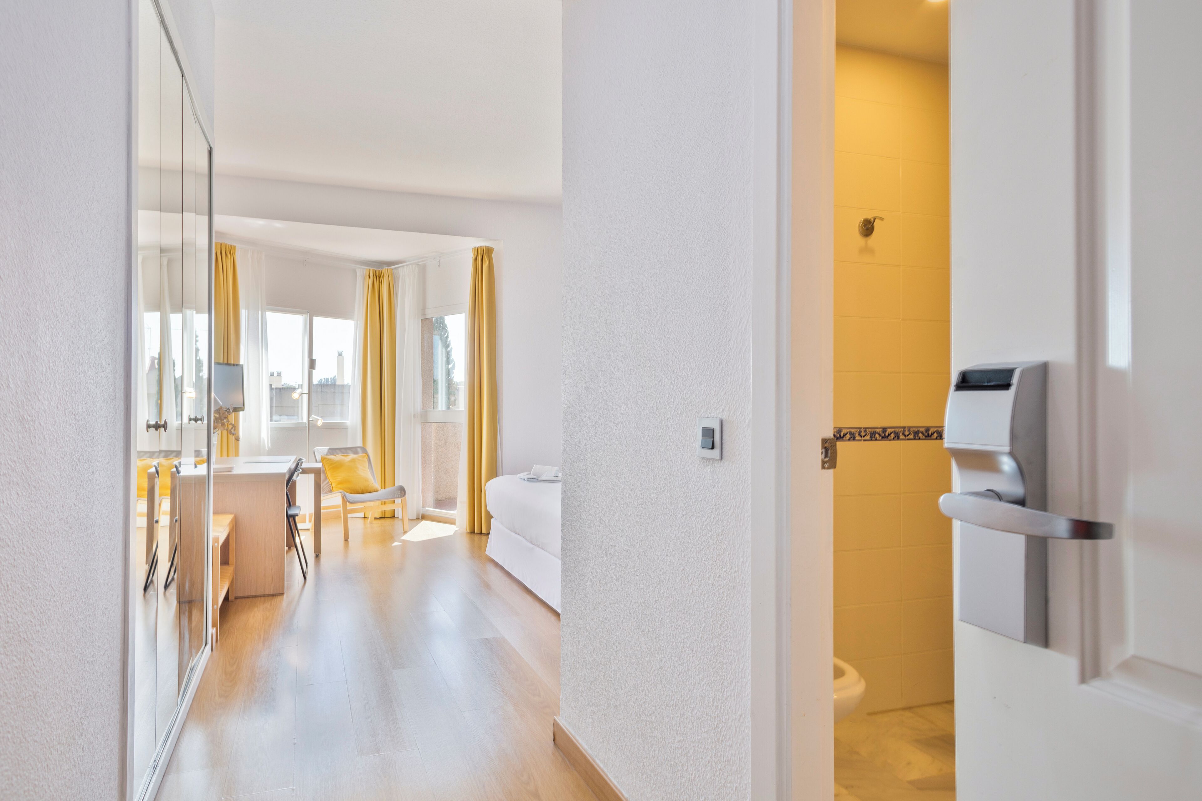 Un pasillo de hotel con suelo de madera, una zona de estar con una silla amarilla, una cama y un baño con inodoro y dispensador de toallas.