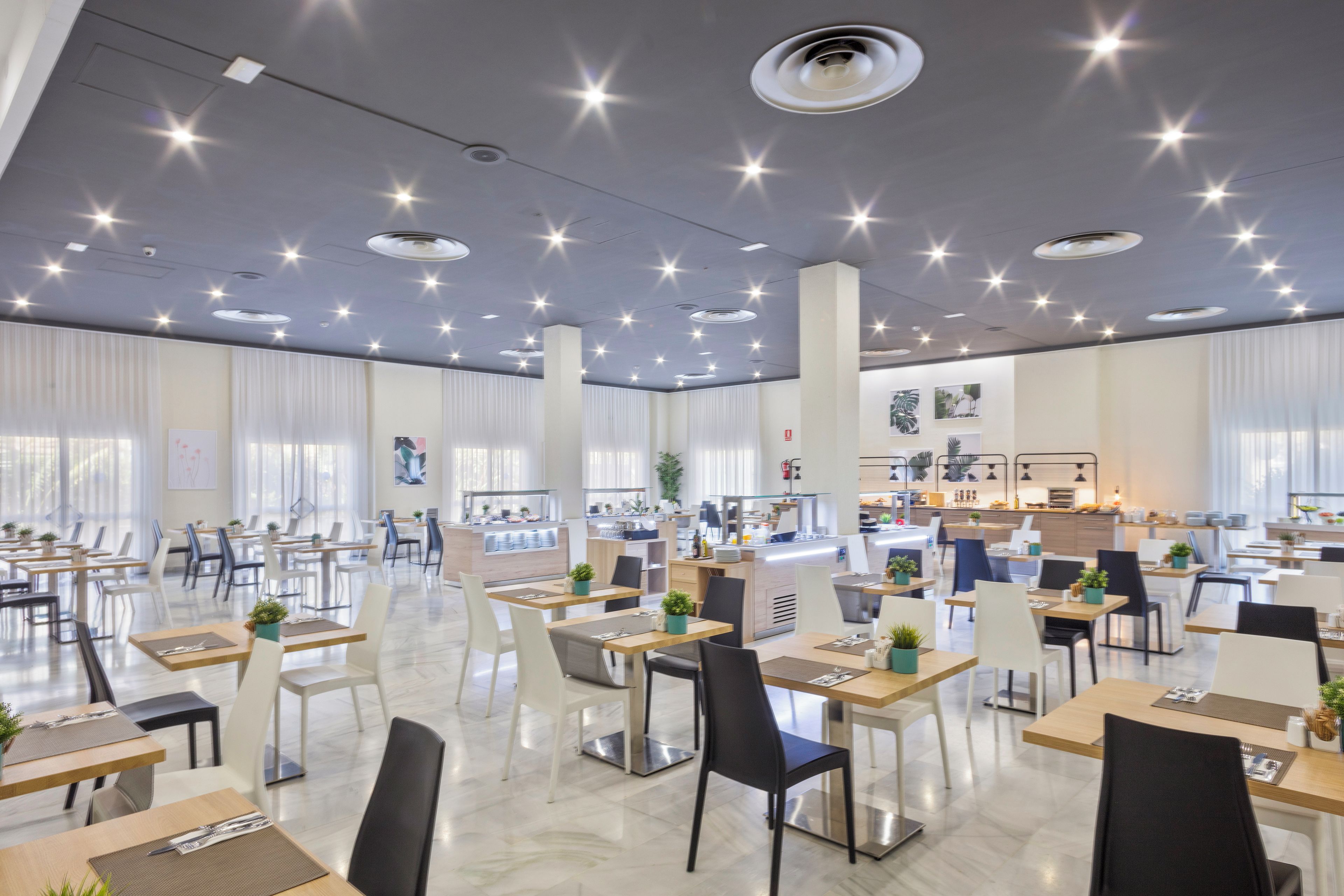Un comedor amplio con múltiples mesas y sillas, un mostrador central de servicio y una cocina al fondo.