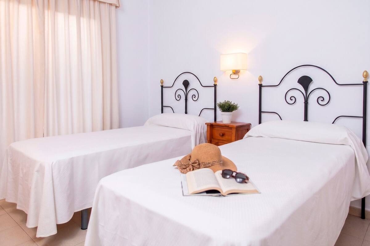 Une chambre d’hôtel avec deux lits simples, une table de chevet avec une lampe, une petite plante en pot et une fenêtre avec des rideaux.