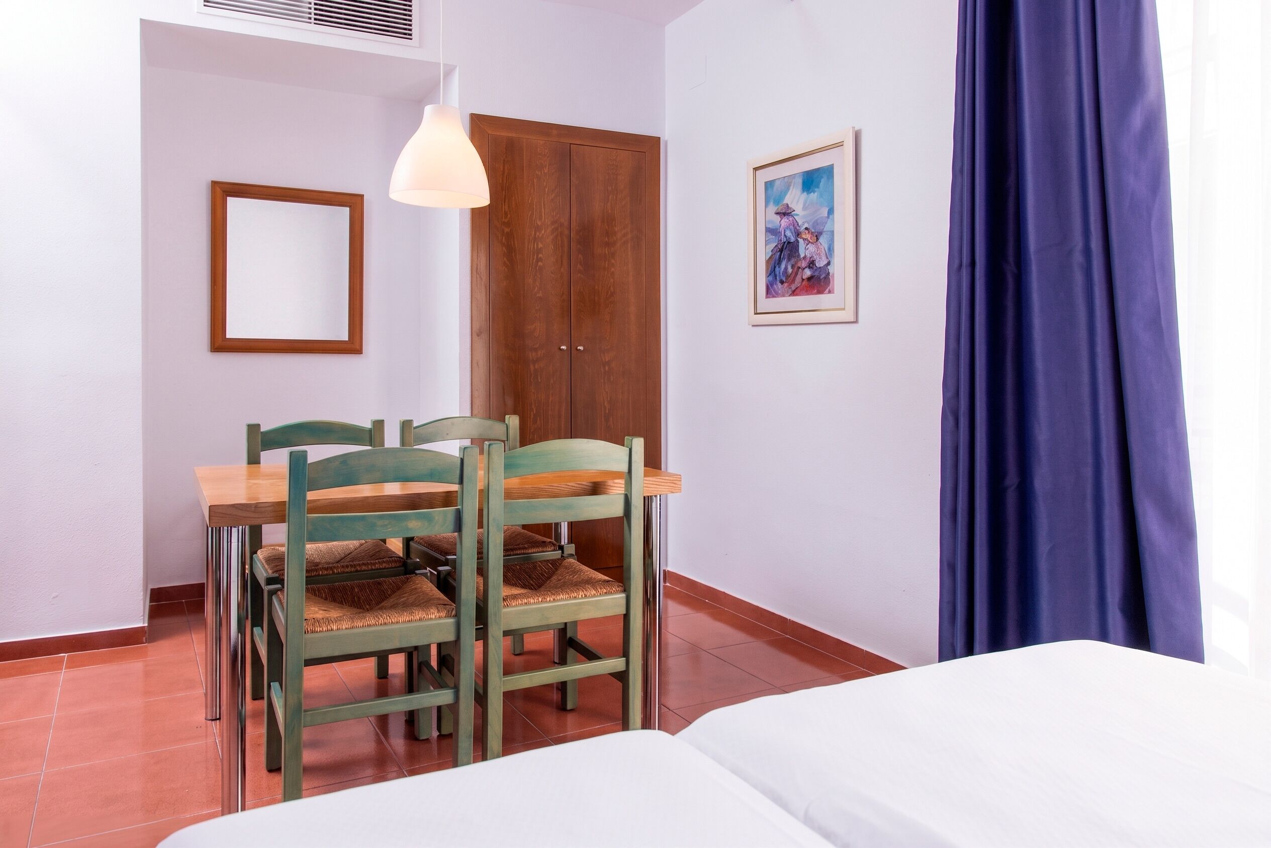 Um quarto com uma cama, uma mesa de jantar de madeira com cadeiras, um lustre pendurado, uma porta e um quadro na parede.