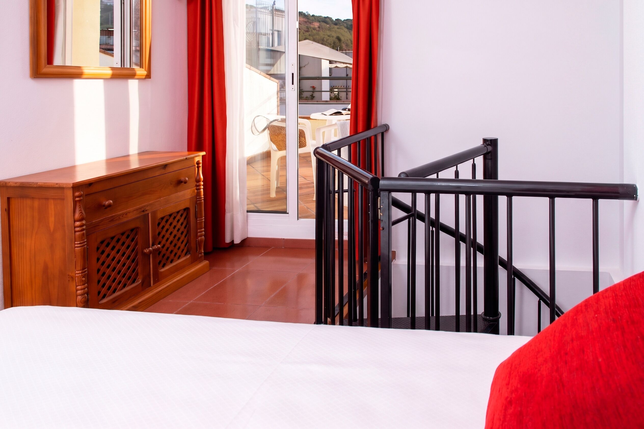 A hotel room with a wooden dresser, a bed, a balcony with red curtains, and a staircase with black railings.