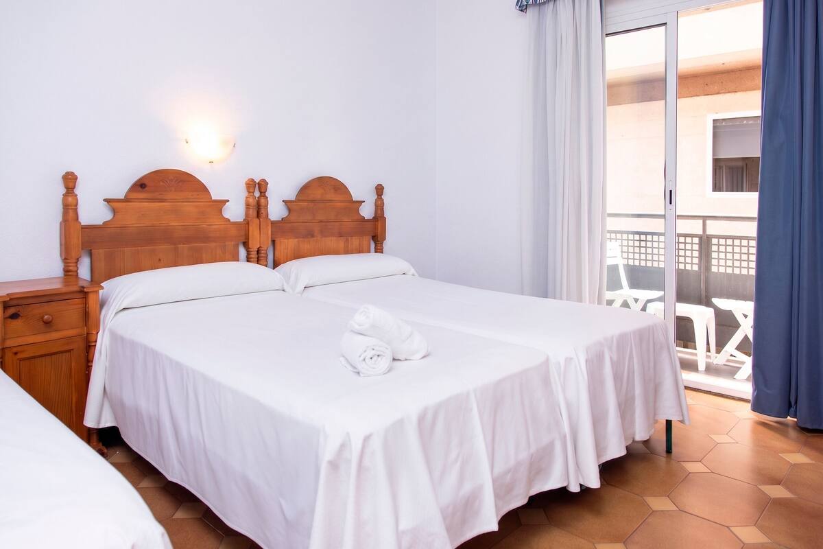 Une chambre d’hôtel avec deux lits, une tête de lit en bois, une table de chevet et un balcon avec des chaises blanches.