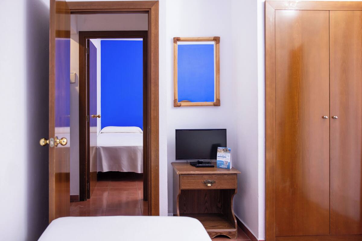 Une chambre d’hôtel comprenant un lit, une table de chevet en bois avec une télévision et une télécommande, une armoire et une porte donnant sur une autre pièce.