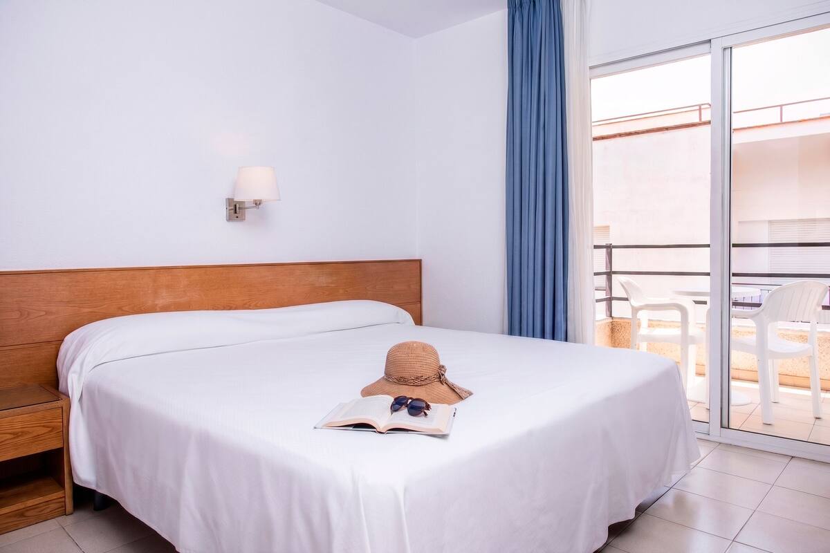 Une chambre d’hôtel comprenant un lit, une table de chevet, une lampe de lecture, une fenêtre donnant sur l’extérieur et un balcon avec des chaises.