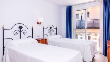 Habitación de hotel con dos camas, una mesita de noche, una lámpara y un balcón con cortinas azules.