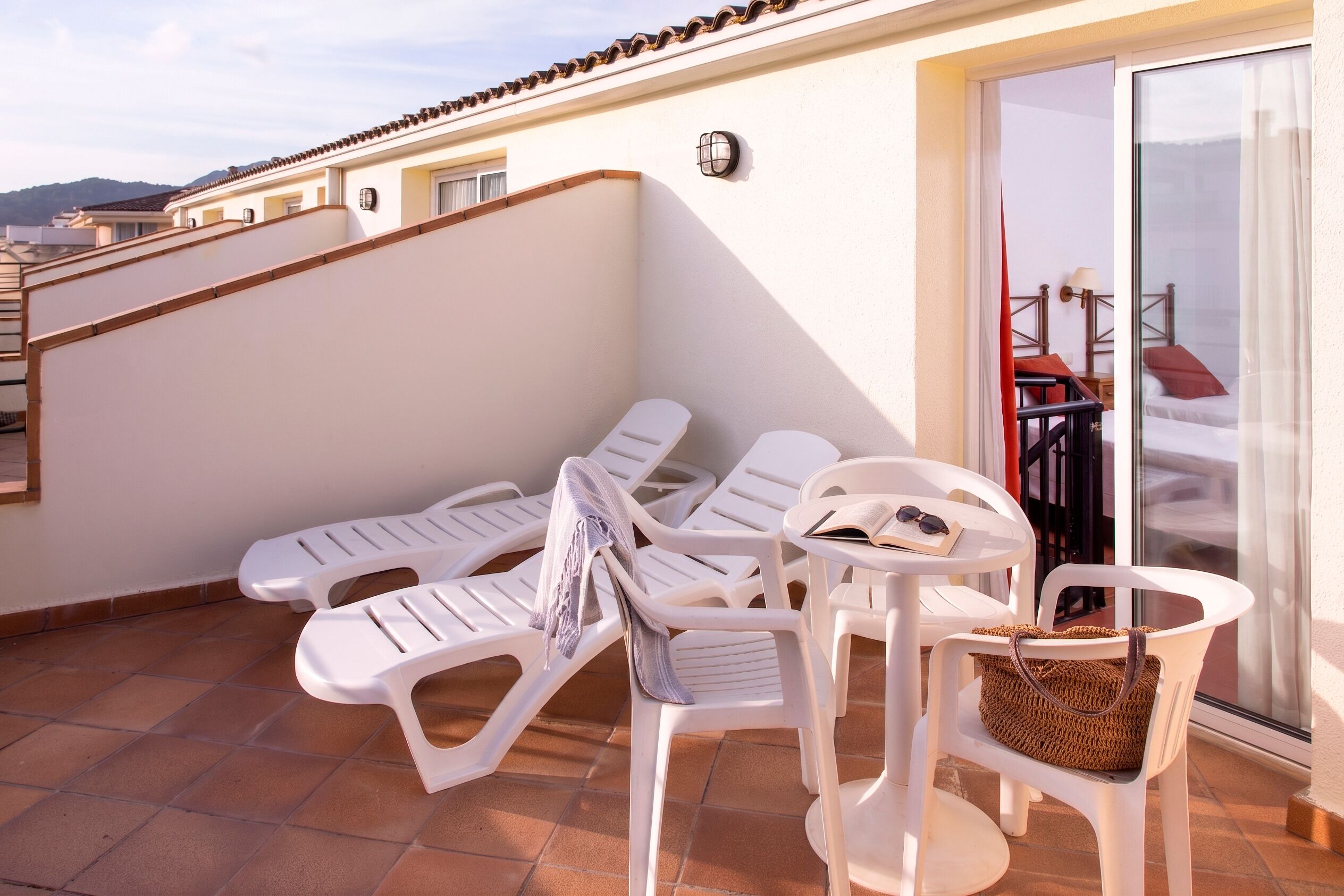 Un balcon avec des chaises en plastique blanc, un panier en osier et une porte-fenêtre coulissante donnant sur une pièce où se trouvent un lit et une table.
