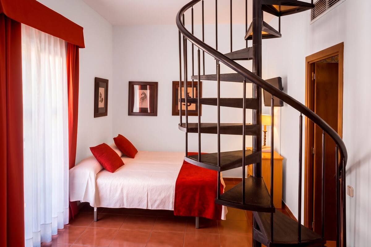 Une chambre à coucher avec un lit, des rideaux rouges, un escalier en colimaçon et des tableaux encadrés accrochés au mur.