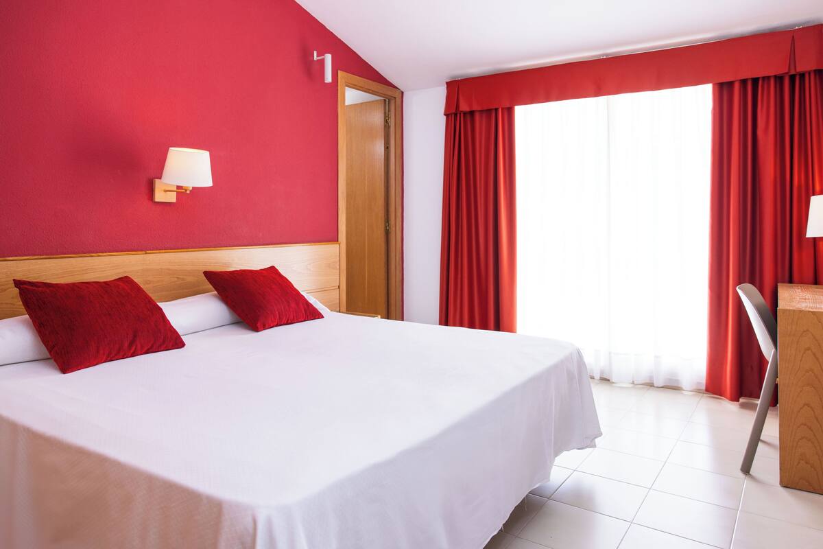 Une chambre d’hôtel avec un grand lit, des rideaux rouges, une tête de lit en bois et un bureau avec une chaise.