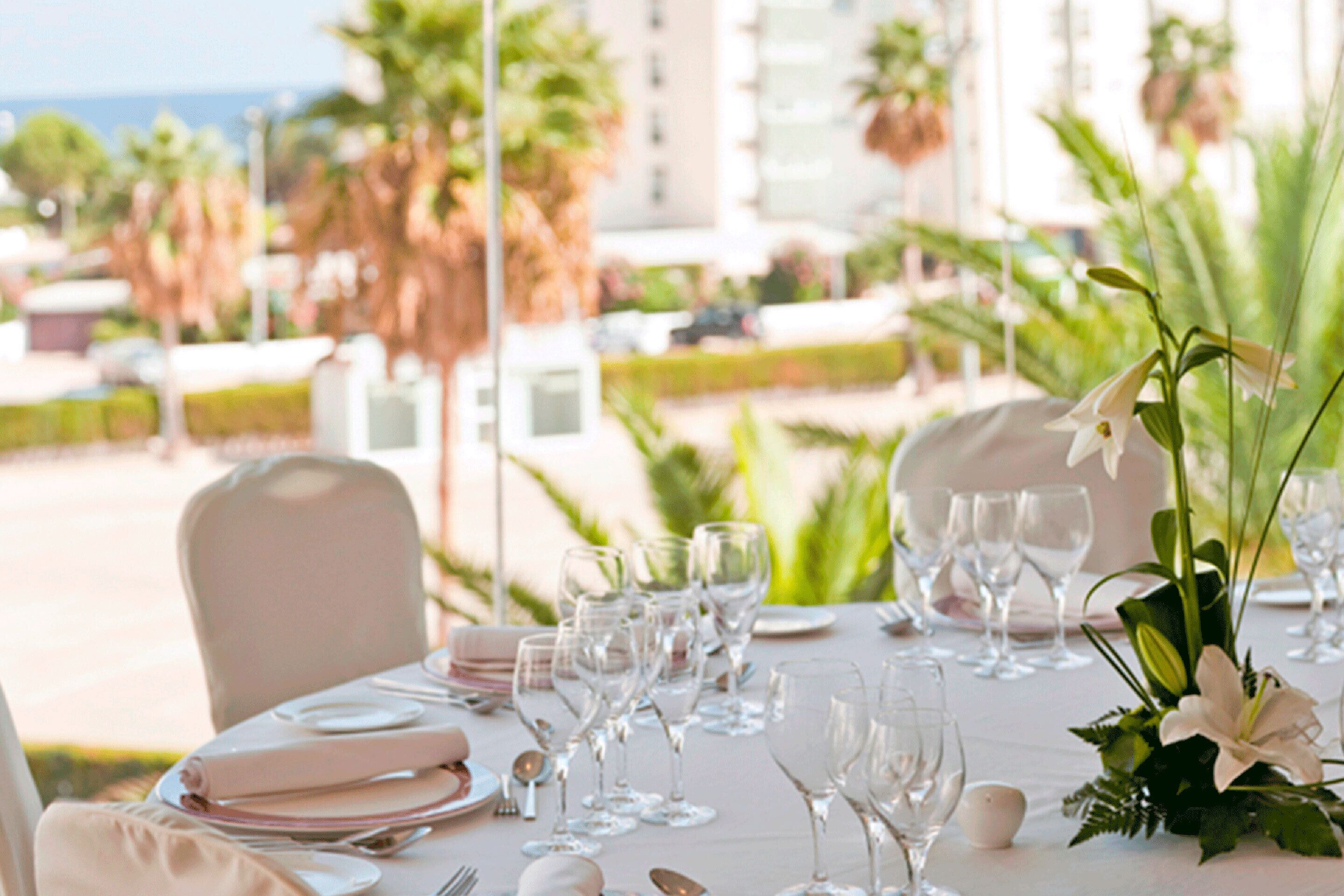 A table set for a formal outdoor dining event with clear glassware and white tableware, surrounded by lush greenery and palm trees, overlooking a scenic view.