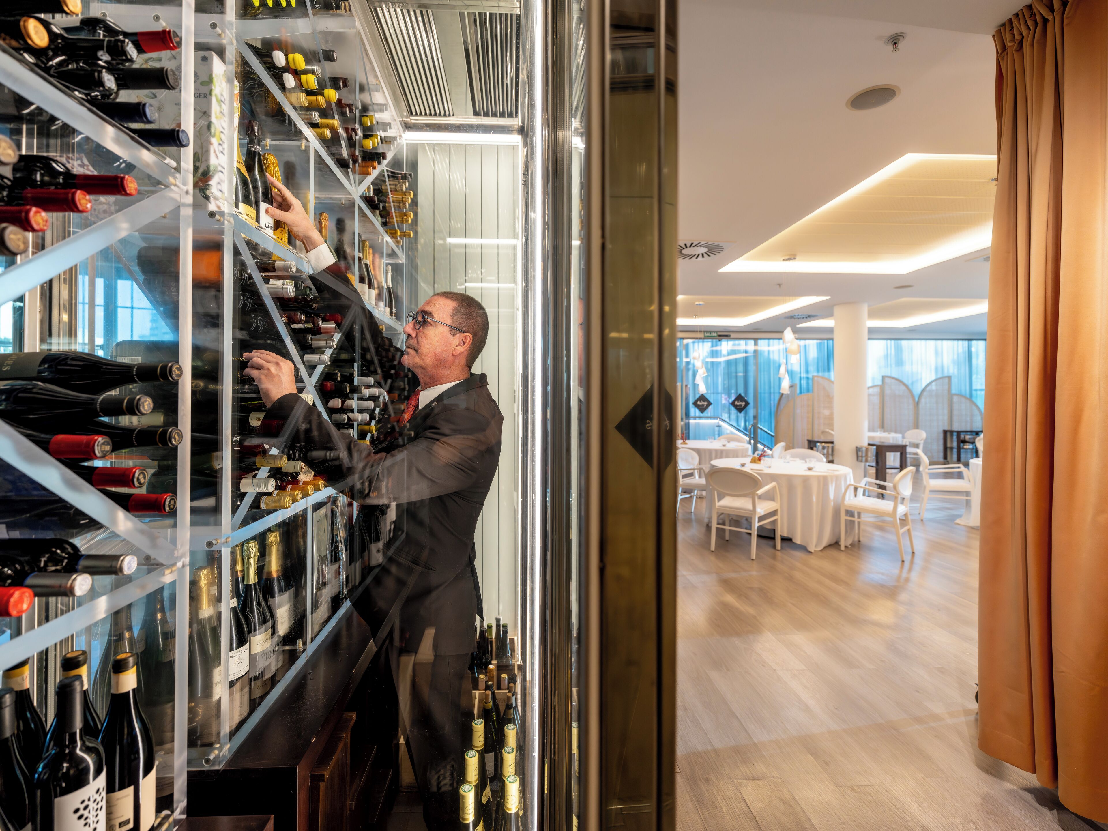 A man organizing wine bottles in a well-lit, modern wine cellar with glass walls and a wooden floor.
