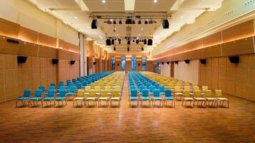 A large hall with rows of blue and yellow chairs, wooden paneling on the walls, and a stage with lighting equipment.