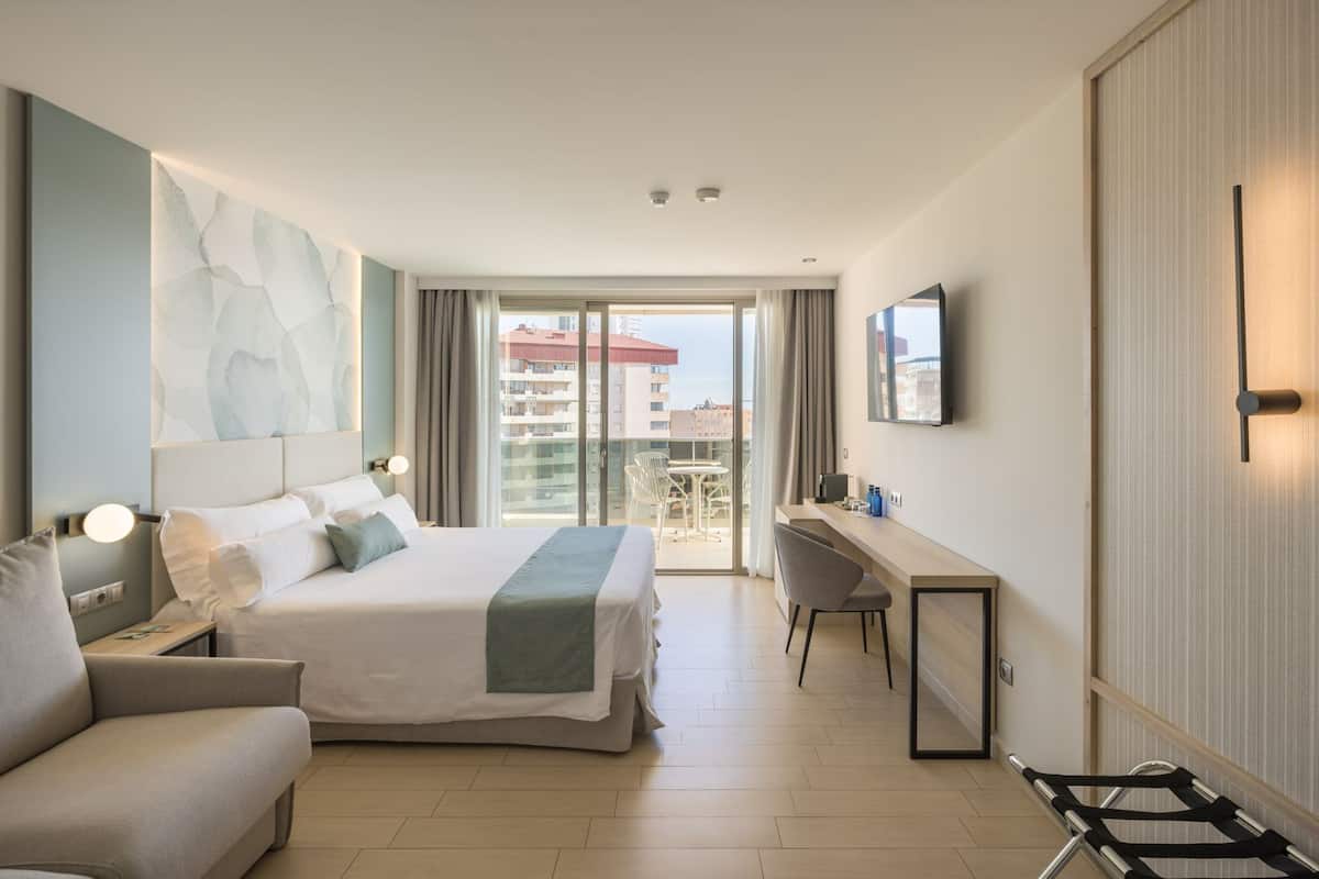 Ein modernes Hotelzimmer mit einem großen Bett, einem Schreibtisch, einem Stuhl und Blick auf die Stadt durch die Balkontüren.