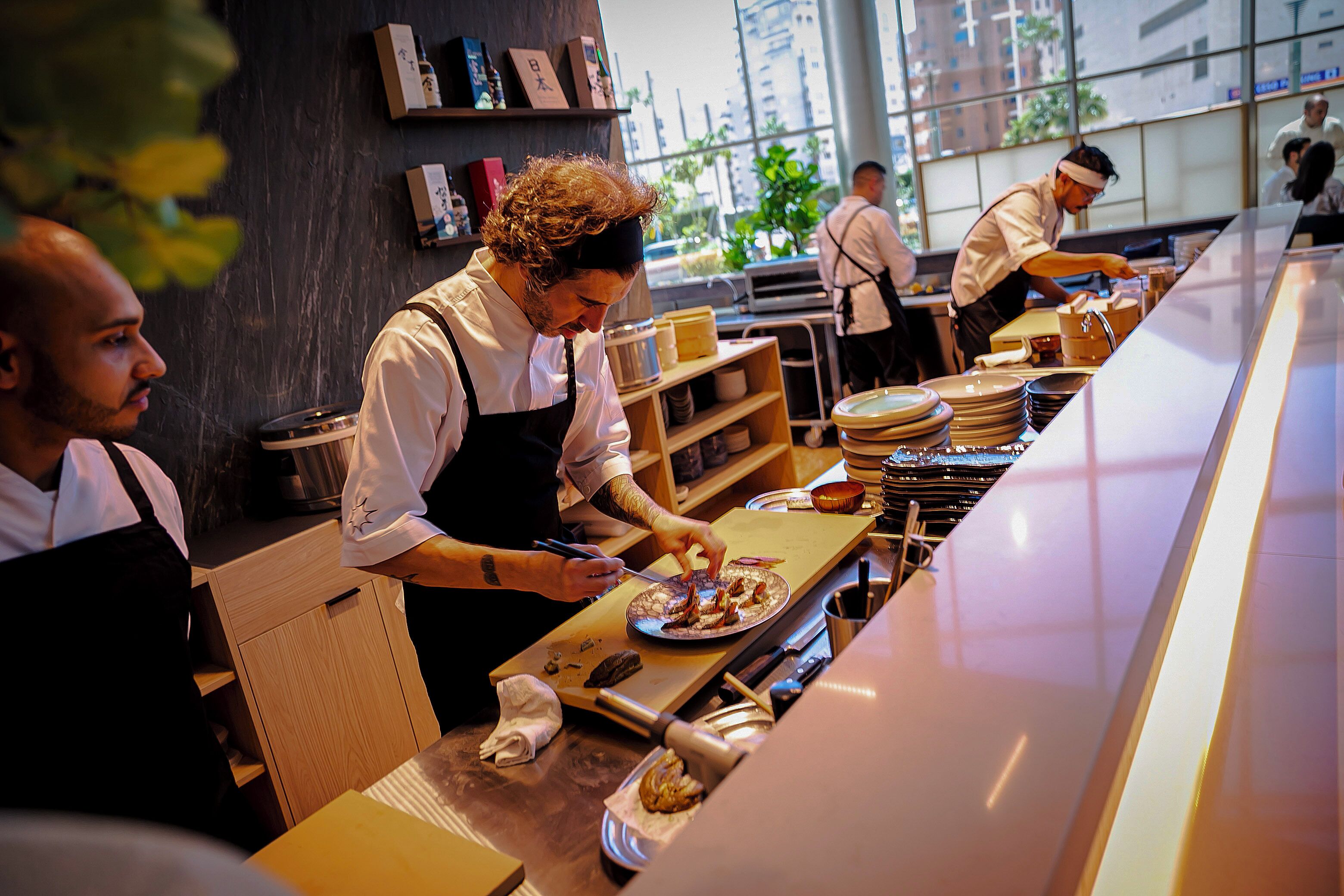 A kitchen area with chefs preparing food, wooden shelves with kitchen items, and a large window providing natural light.