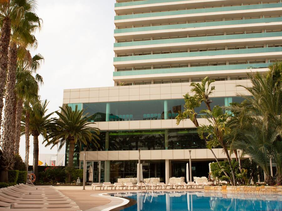 Un hôtel doté d’une piscine, de chaises longues, de palmiers et d’un bâtiment moderne aux lignes horizontales.