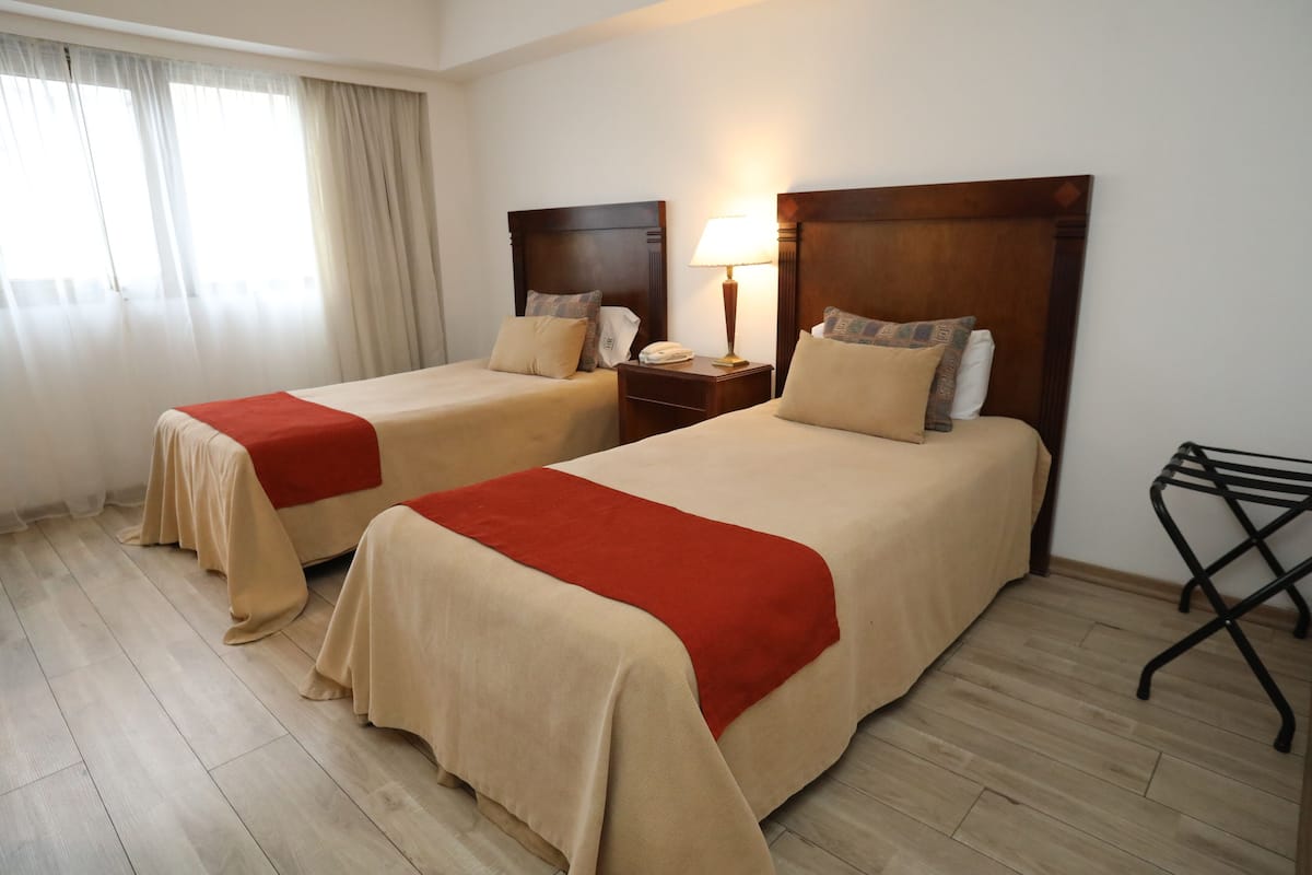 A hotel room with two beds, wooden headboards, beige bedding, a red throw blanket, a nightstand with a lamp, and a black folding chair.