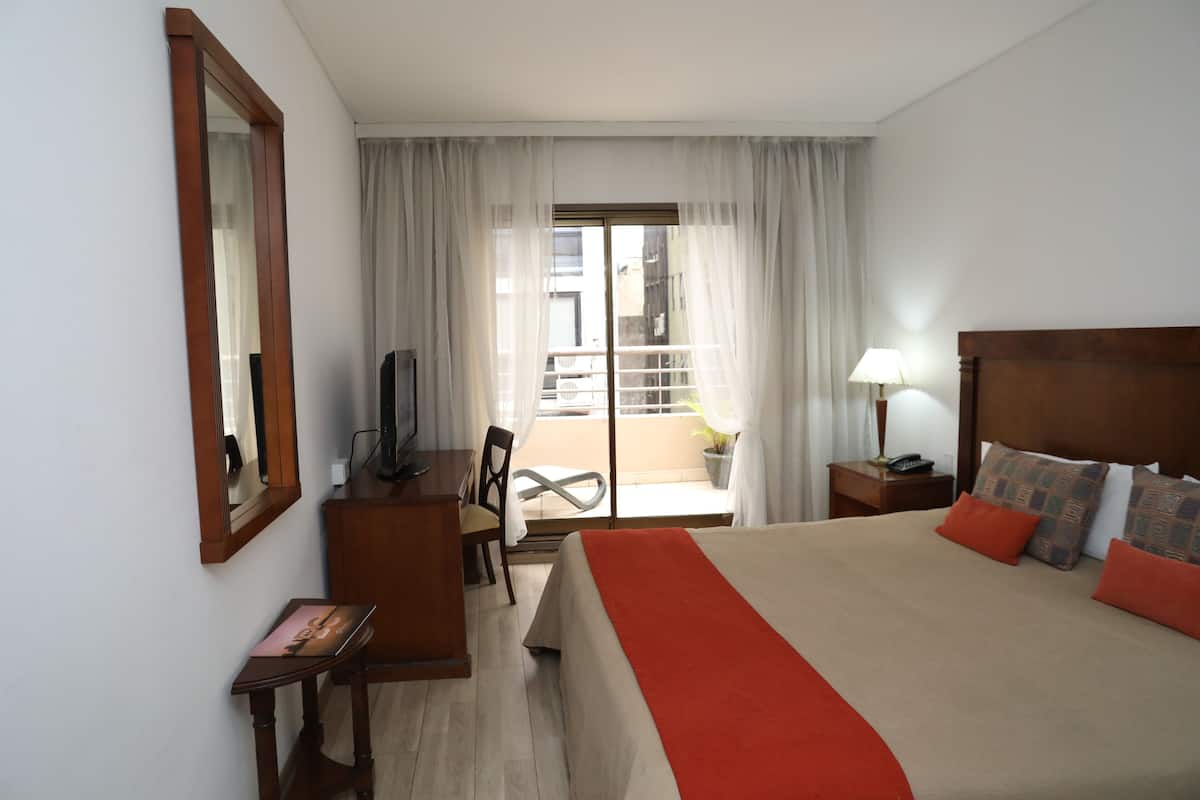 A hotel room with a bed, a desk, a chair, a TV, a nightstand, and a window with curtains.
