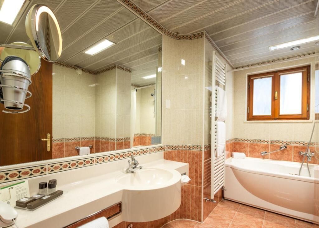 A bathroom with a double sink, a large mirror, a bathtub, and a window.